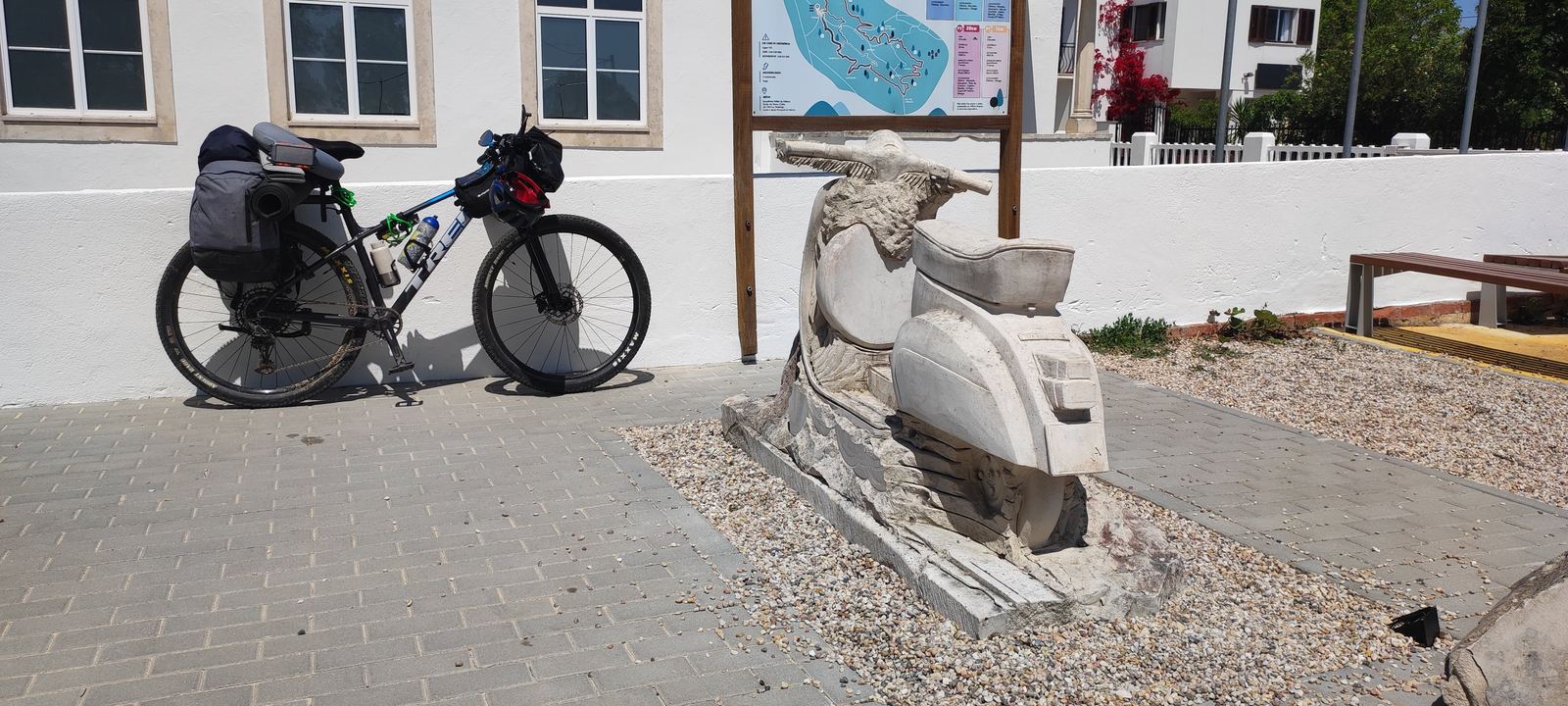 A loaded bike with a sleeping bag on the rack, parked against a white wall beside a panel showing local route distances