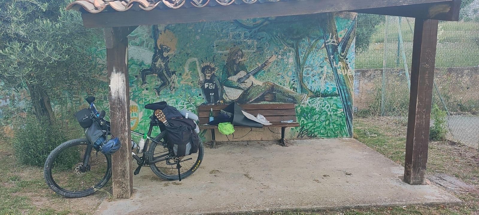 A loaded bike leaning against the pillar of a roadside shelter, bags on a wooden bench, a mural painted on the wall behind
