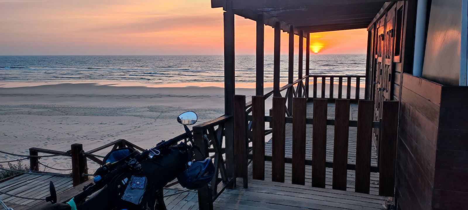 The handlebars of a loaded bike, helmet hanging from the bar bag, stopped on a wooden boardwalk above a wide empty beach at dusk