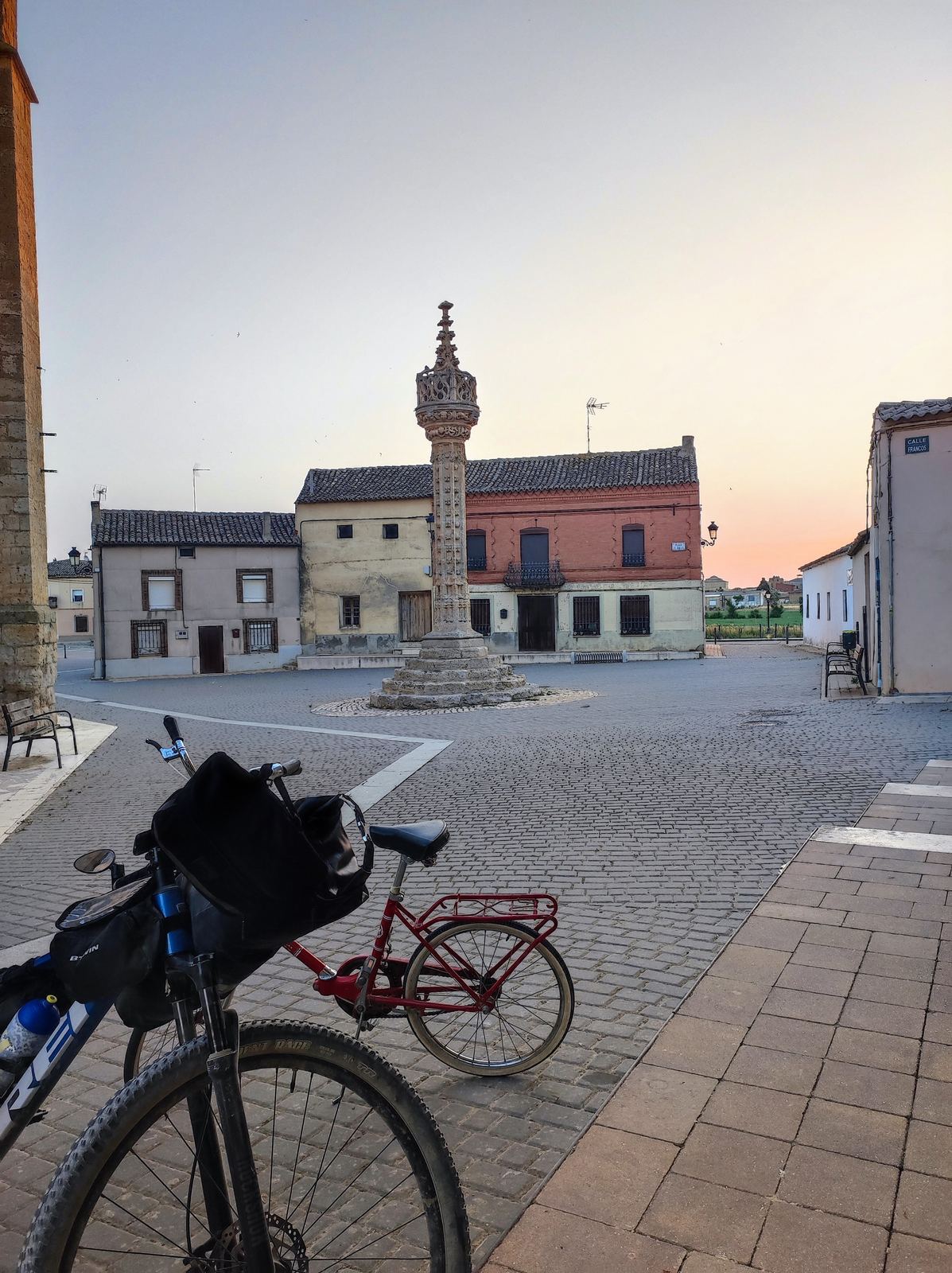 A loaded bike in the foreground of an empty paved square at dawn, a gothic stone column at its centre, low brick houses and a pink sky