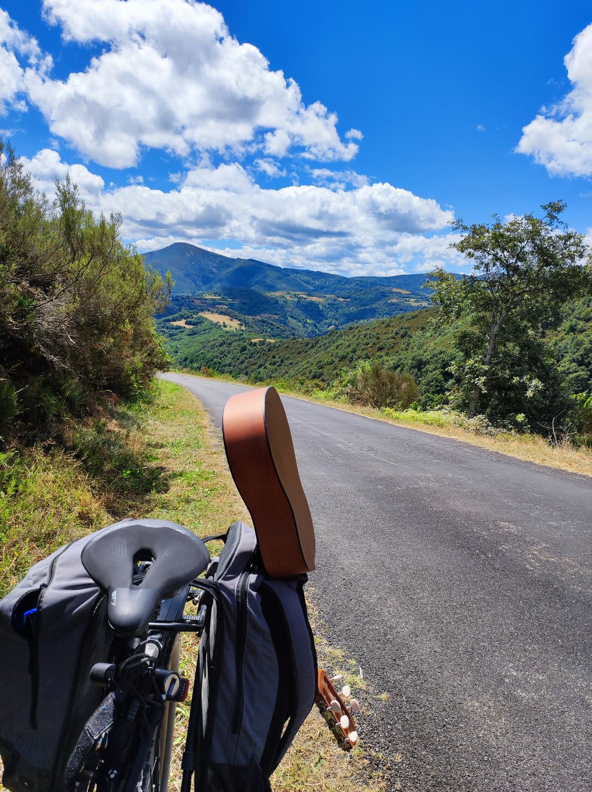 Seen from behind a loaded bike's saddle and bags: an asphalt road dropping away towards a valley of green mountains under high cloud
