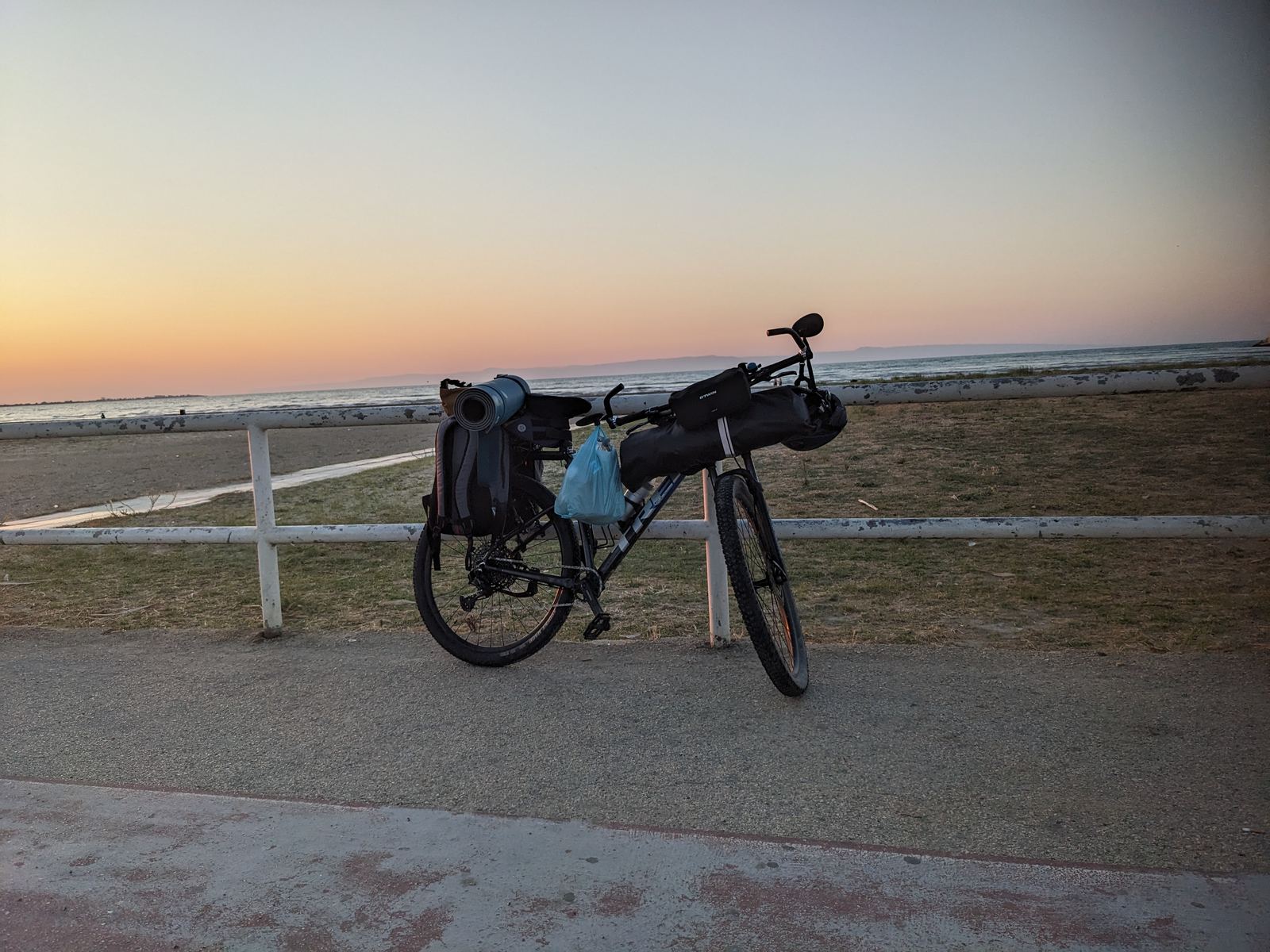 A touring bike loaded with a rolled sleeping mat, rucksack and handlebar bag, leaning on a white fence above an empty beach at sunset