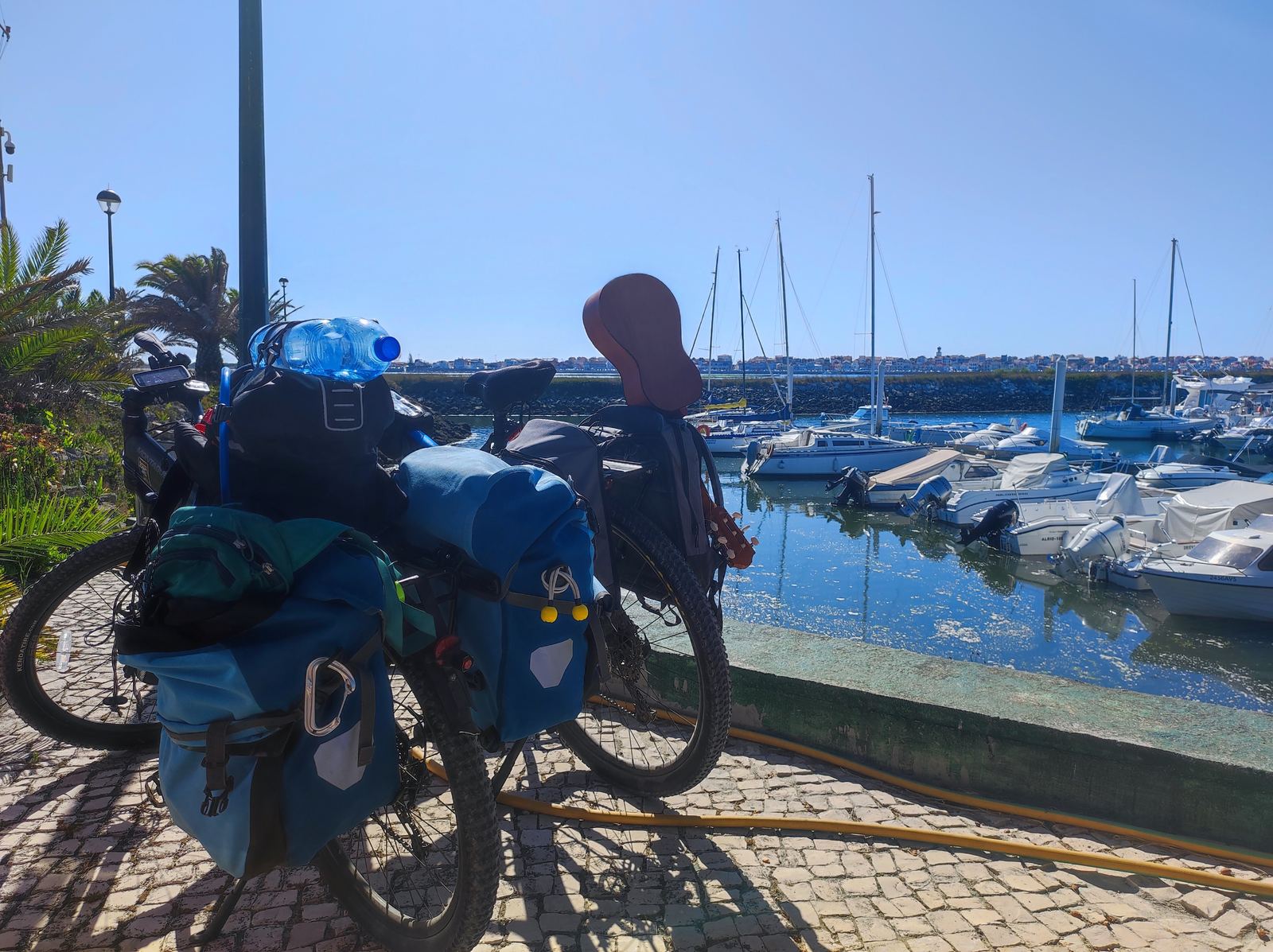 A bike with blue front and rear panniers and a green dry bag on the rack, standing on the cobbles of a small harbour