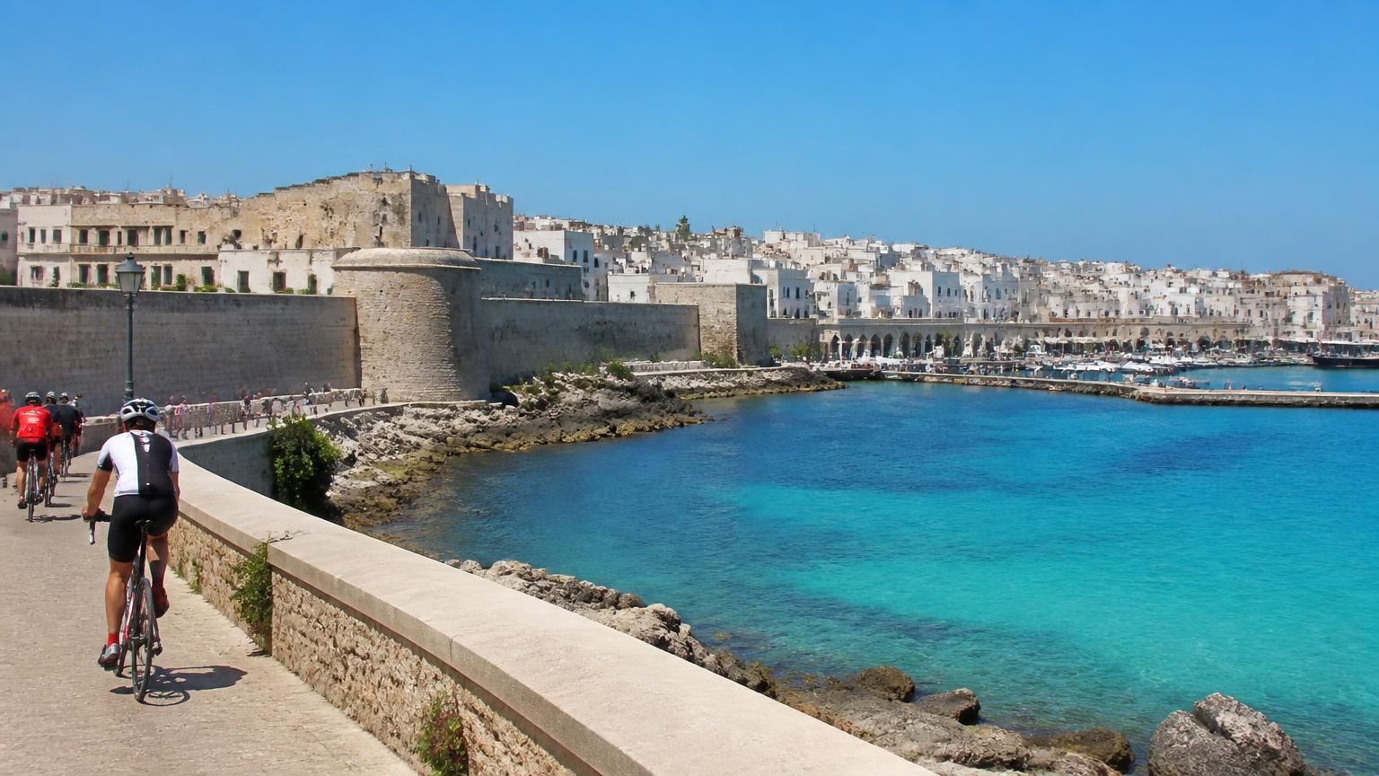 Road cyclists riding along the seafront wall at Otranto, the white town and its harbour behind them