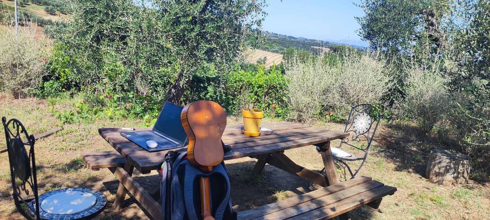 A wooden picnic table under olive trees with an open laptop, a yellow mug, a travel rucksack and a guitar leaning against it
